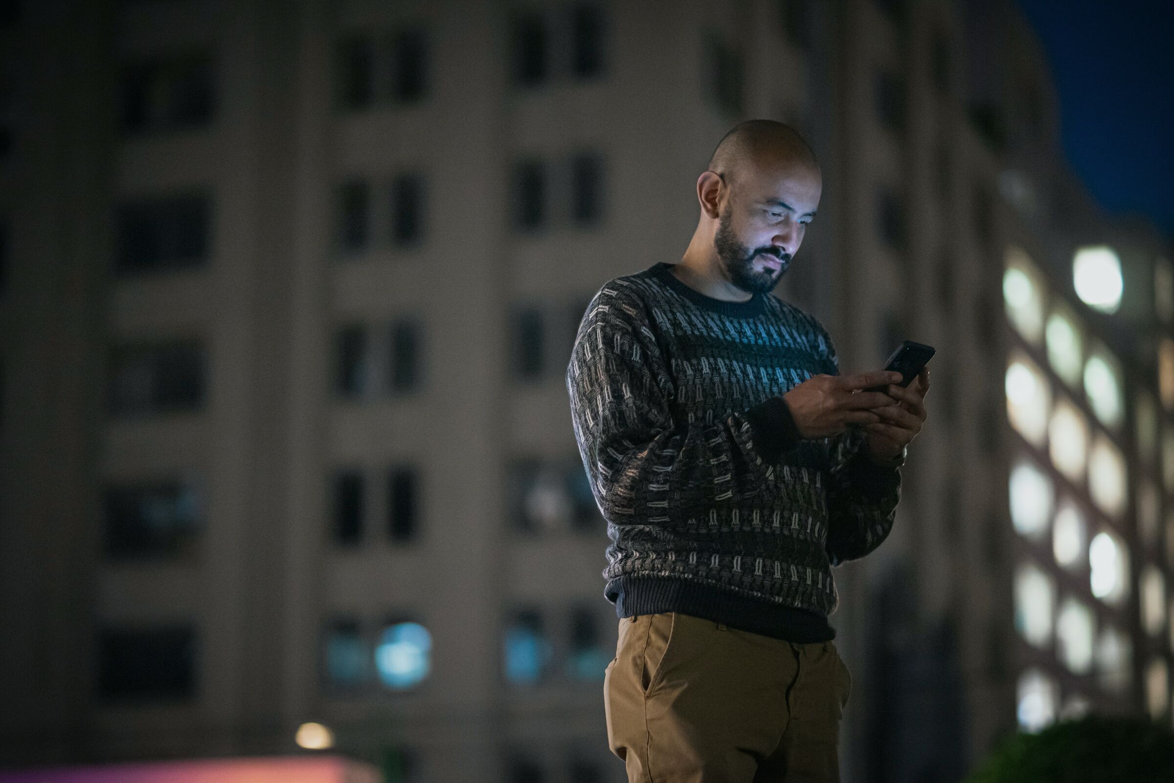 A man standing in a city at night, looking down at his phone