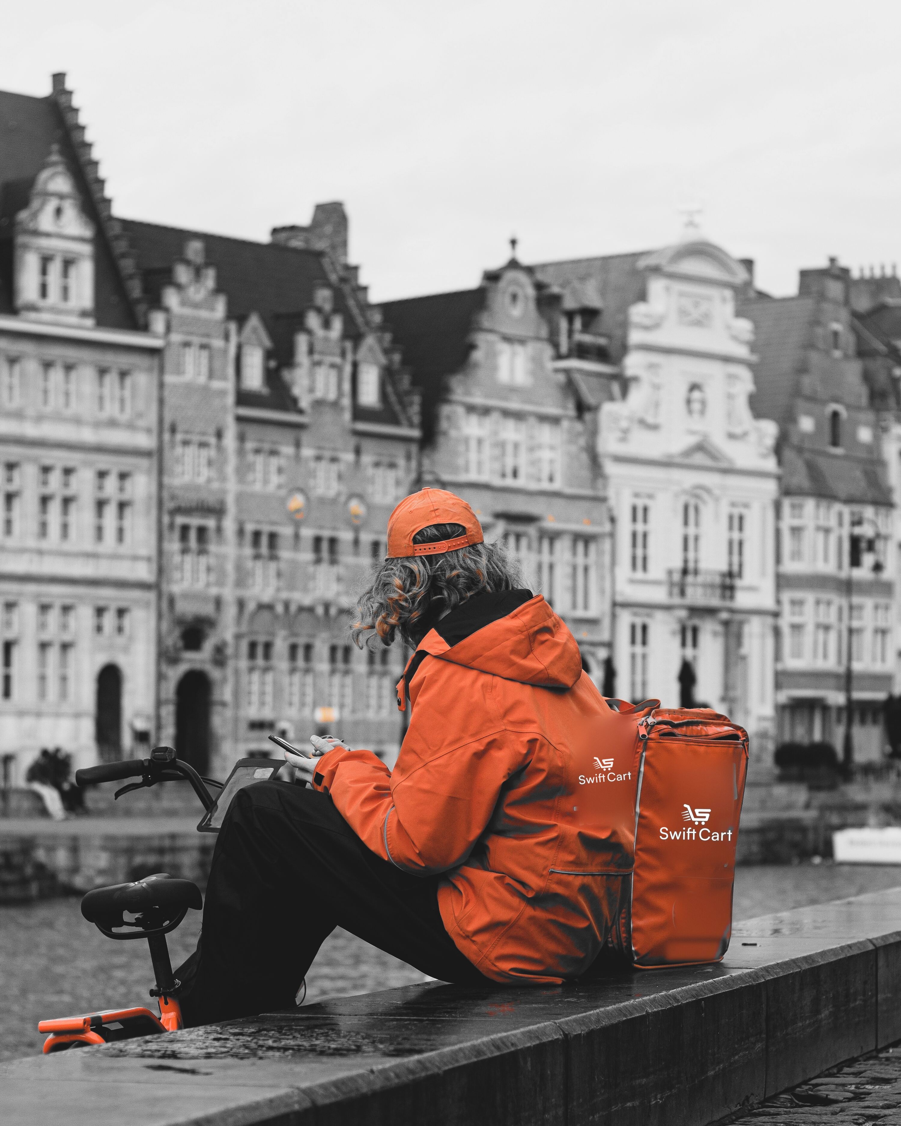 A SwiftCart courier in an orange jacket sitting on a canal wall, checking a phone beside the delivery bag